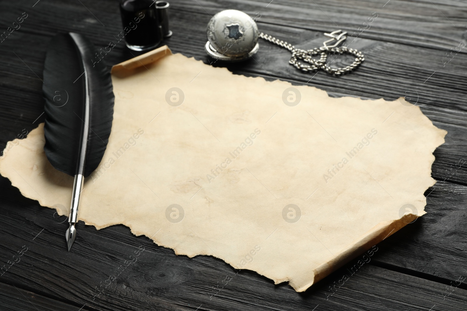 Photo of Sheet of old parchment paper, feather, inkwell and pocket chain clock on black wooden table