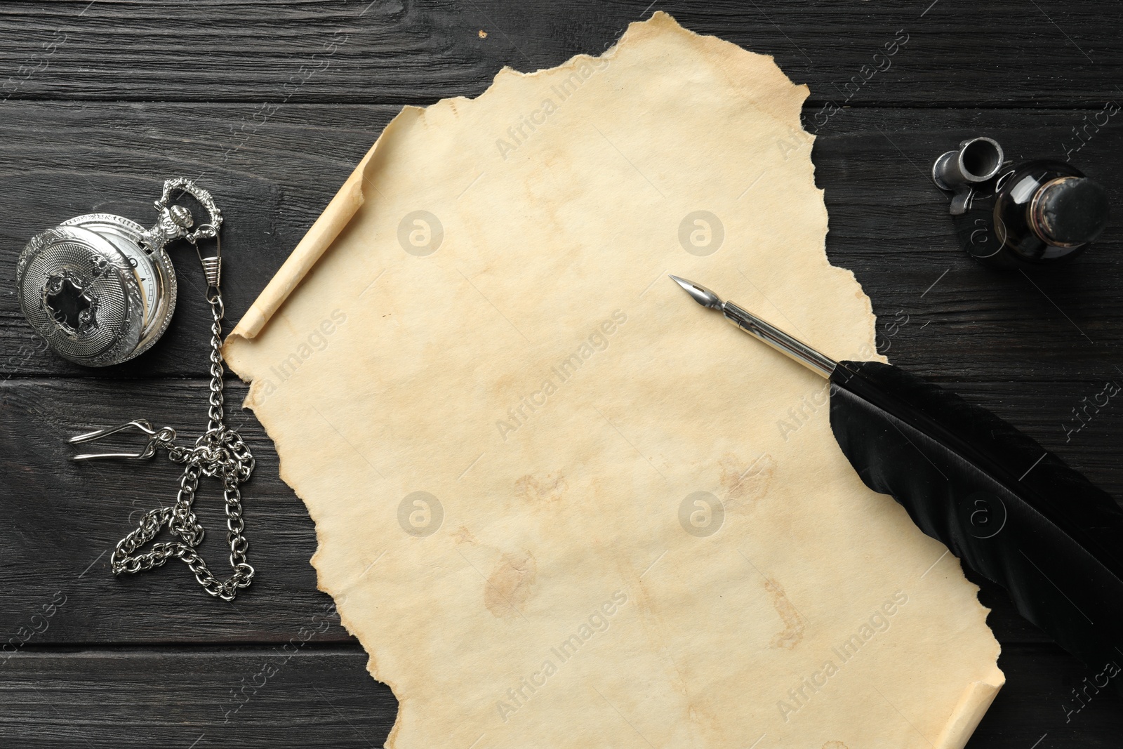 Photo of Sheet of old parchment paper, feather, inkwell and pocket chain clock on black wooden table, top view