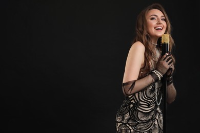 Beautiful young woman in stylish dress with microphone singing on black background, space for text