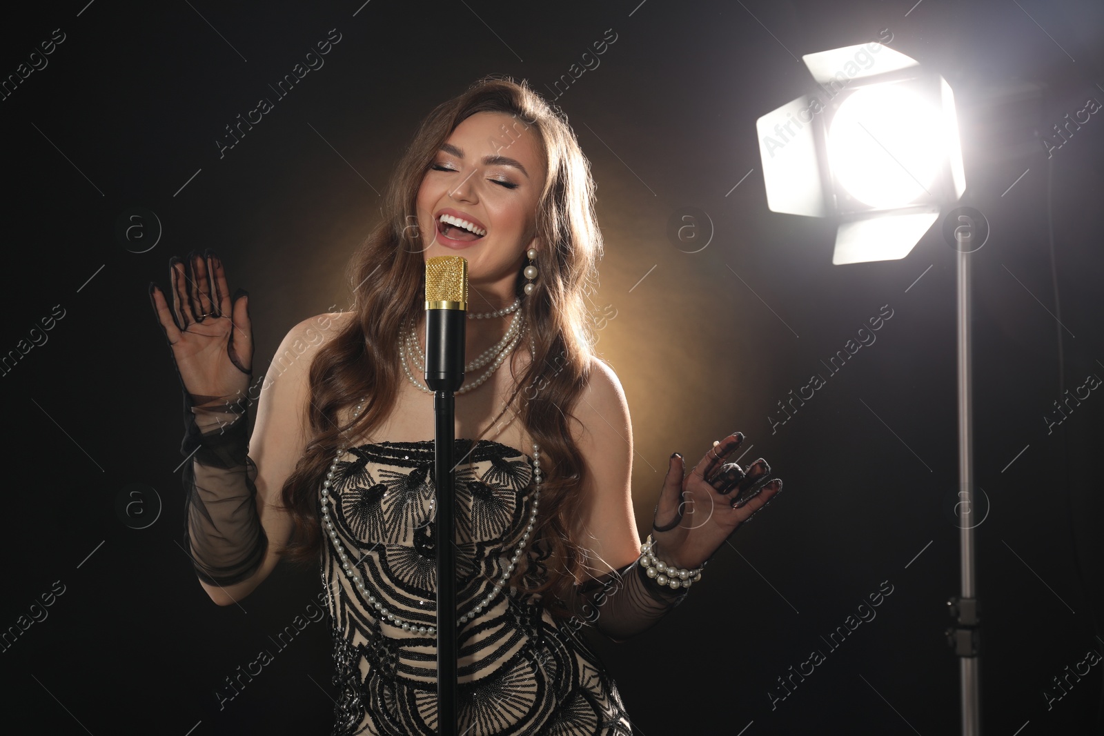 Photo of Beautiful young woman in stylish dress with microphone singing on dark background