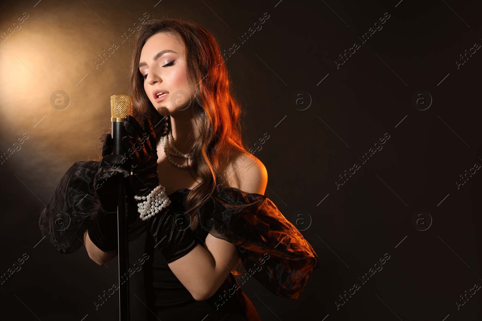 Photo of Beautiful young woman in stylish dress with microphone singing on dark background, space for text