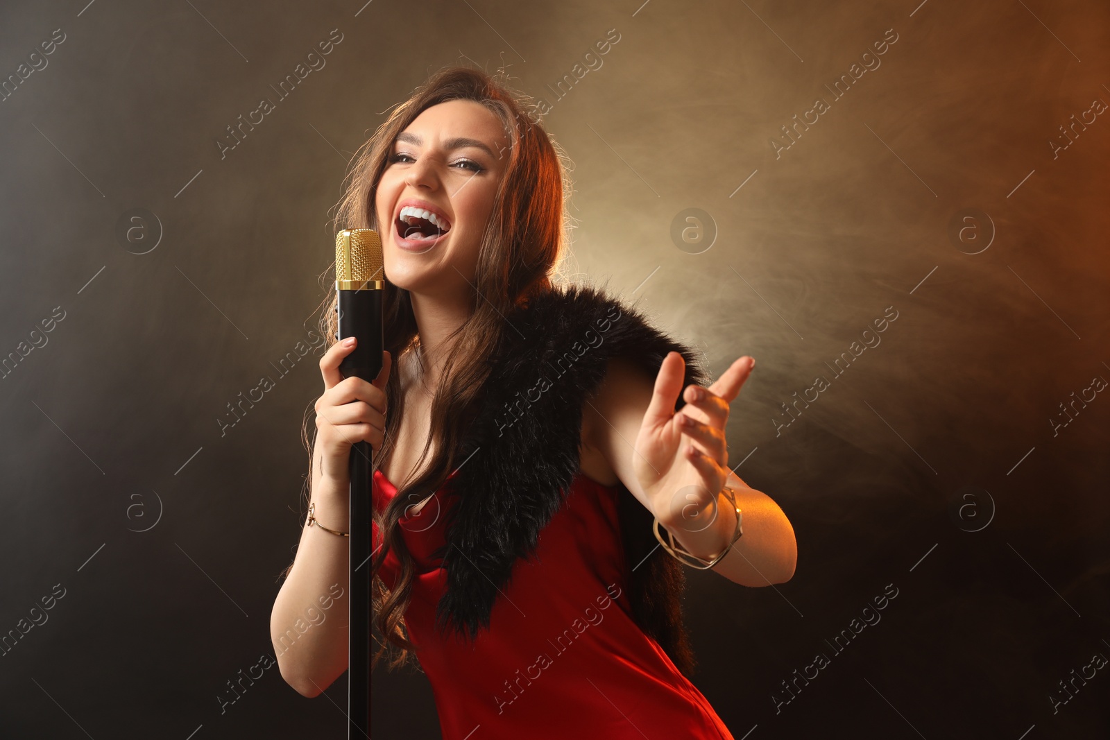 Photo of Beautiful young woman in stylish dress with microphone singing on dark background in color lights and smoke