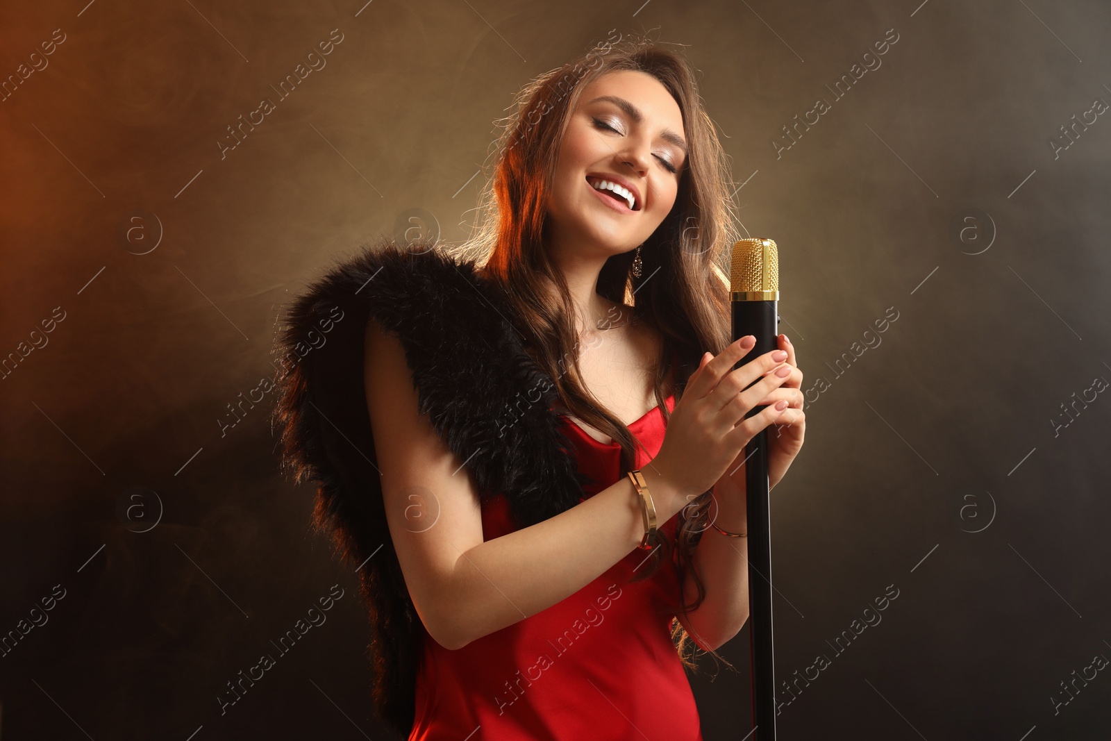 Photo of Beautiful young woman in stylish dress with microphone singing on dark background in color lights and smoke