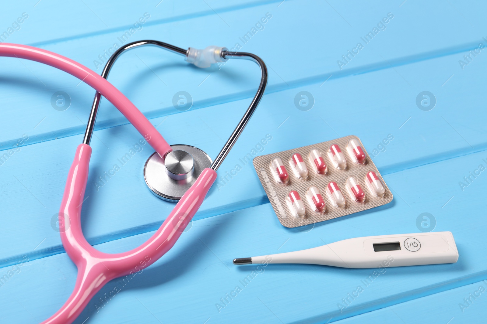 Photo of Stethoscope, thermometer and pills on light blue wooden background, closeup. Medical treatment