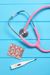 Stethoscope, thermometer and pills on light blue wooden background, flat lay. Medical treatment