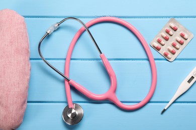 Pillow, stethoscope, thermometer and pills on light blue wooden background, flat lay