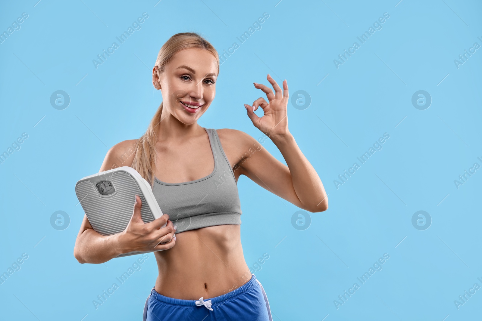 Photo of Happy woman with floor scale showing ok gesture on light blue background. Space for text