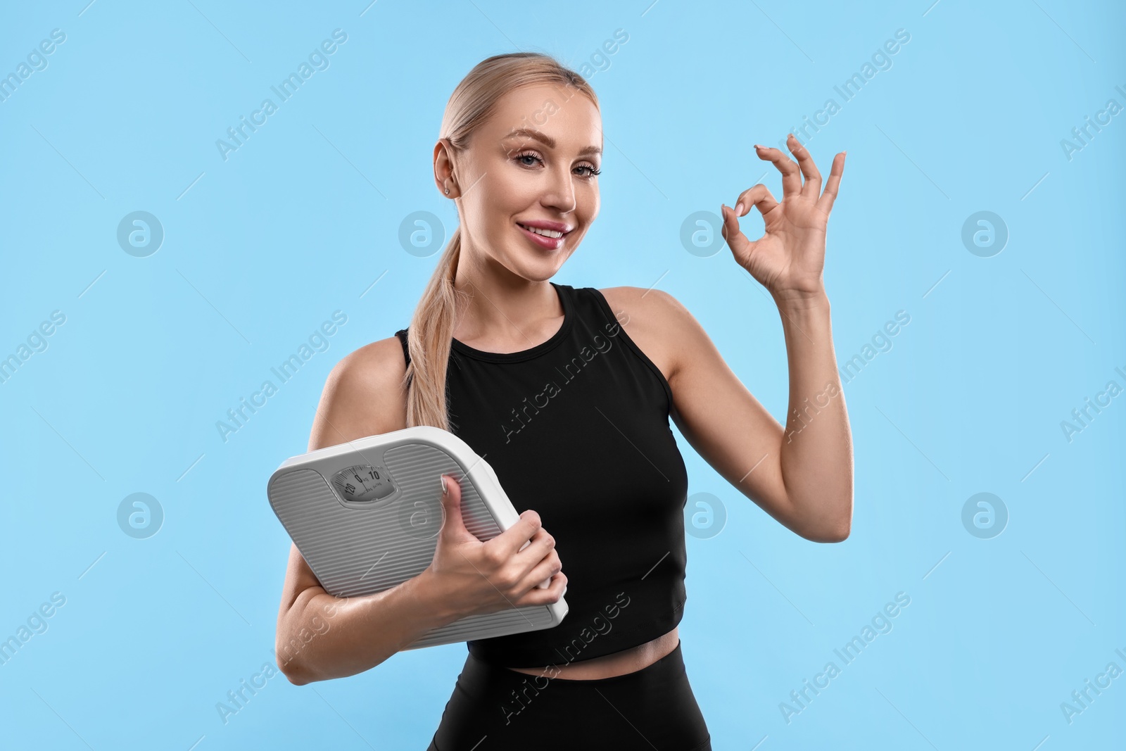 Photo of Happy woman with floor scale showing ok gesture on light blue background