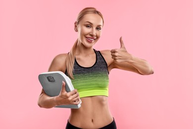 Happy woman with floor scale showing thumb up on pink background