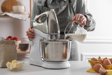Photo of Woman pouring milk into bowl of stand mixer while making dough at table indoors, closeup