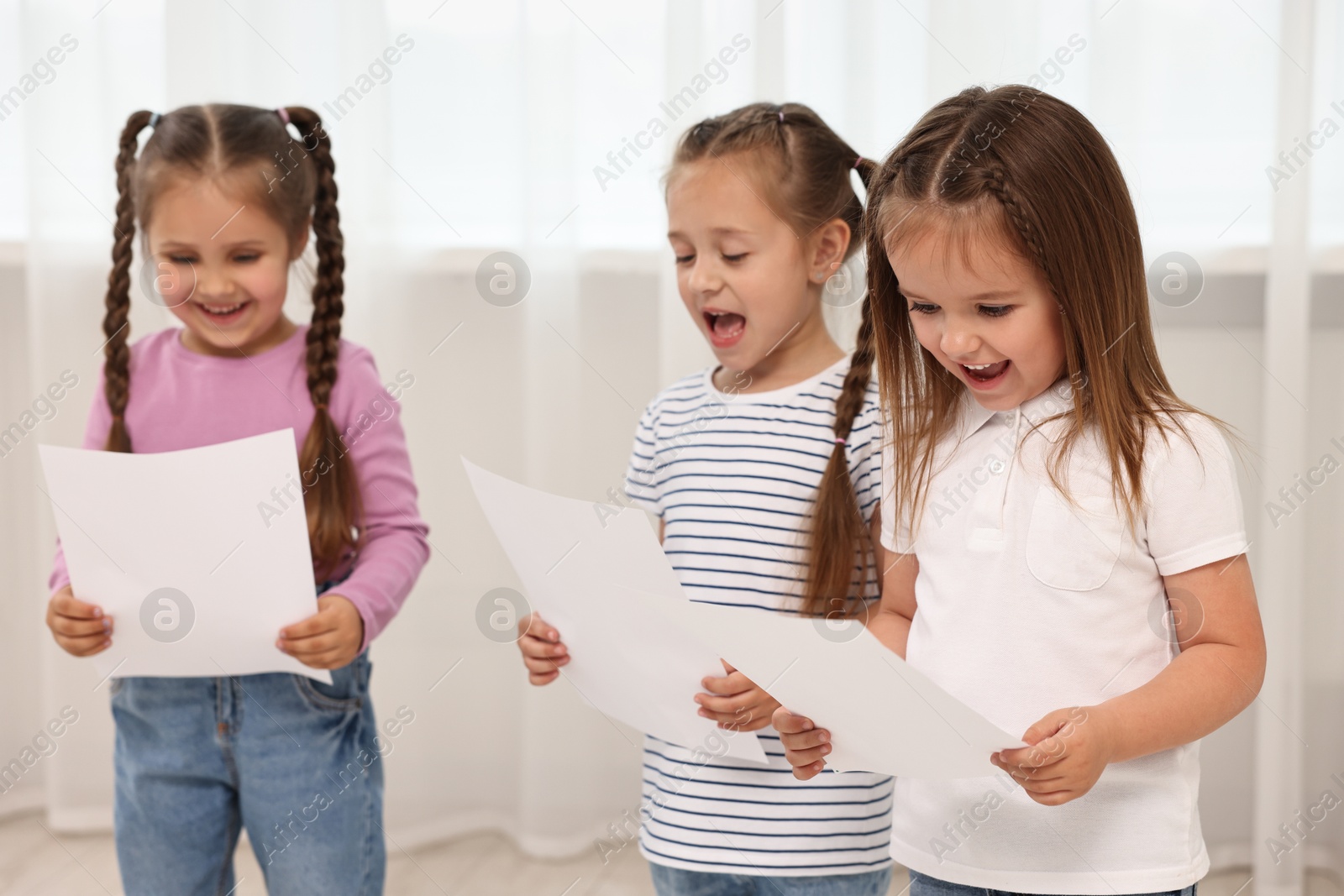 Photo of Cute little children with sheets of paper singing indoors