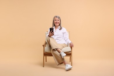 Senior woman with phone on armchair against beige background