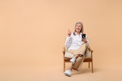 Senior woman with phone on armchair against beige background, space for text