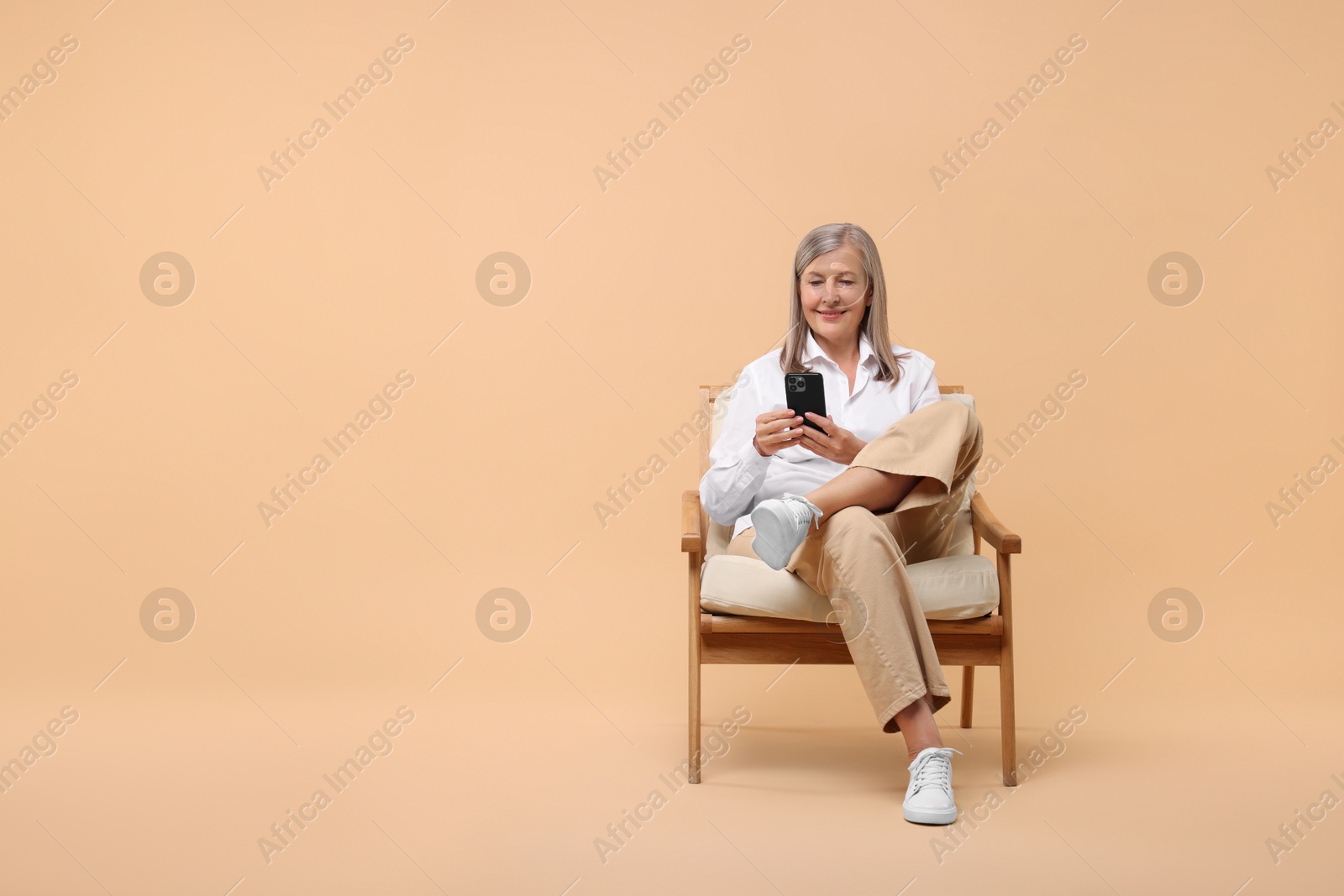 Photo of Senior woman with phone on armchair against beige background, space for text