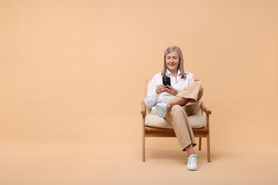 Senior woman with phone on armchair against beige background, space for text