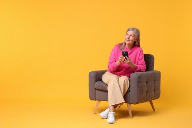 Senior woman with phone on armchair against orange background, space for text