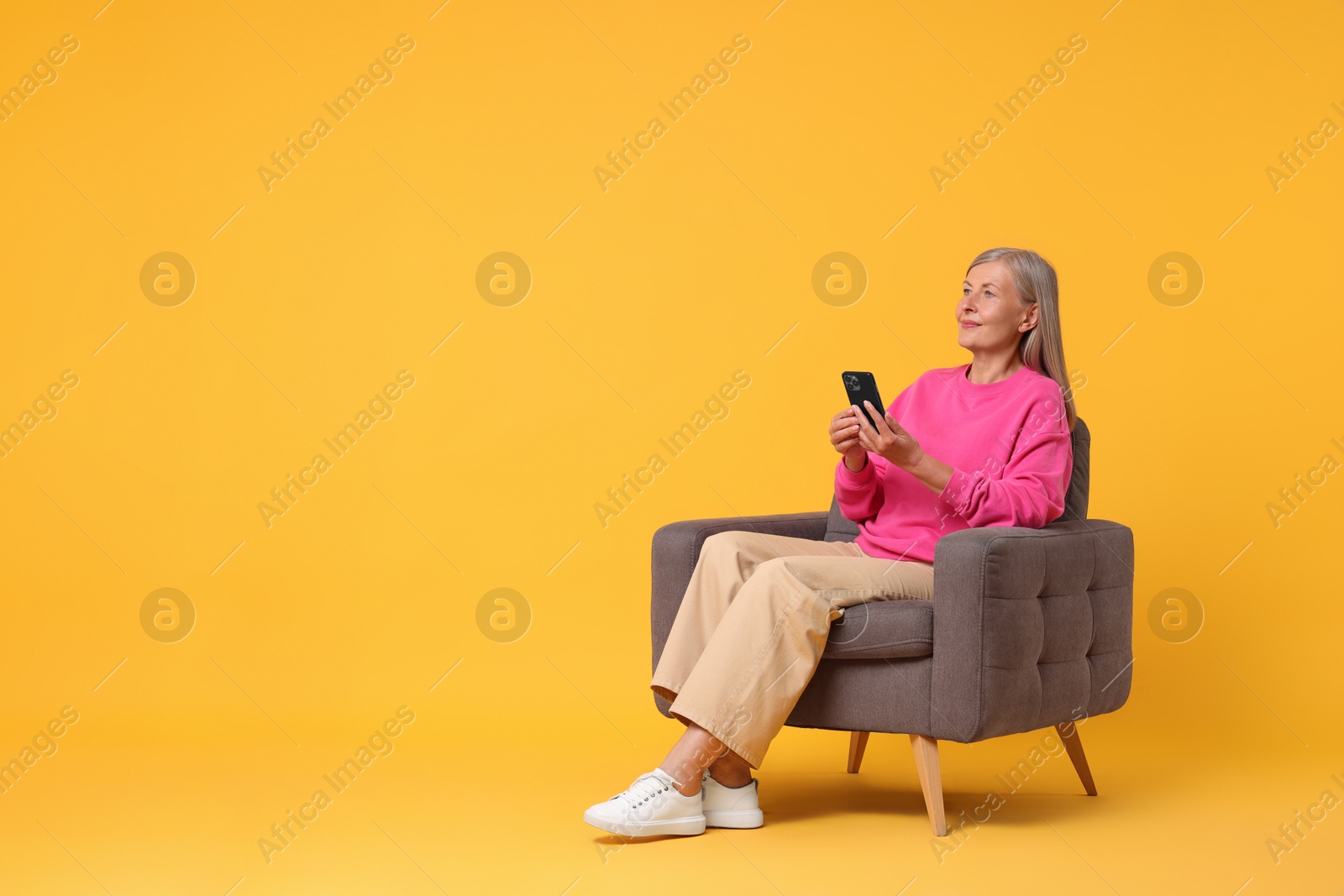 Photo of Senior woman with phone on armchair against orange background, space for text