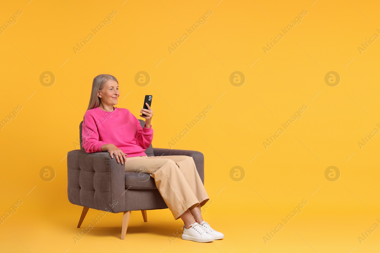 Photo of Senior woman with phone on armchair against orange background, space for text