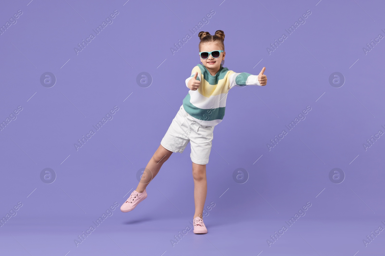 Photo of Cute little girl in sunglasses showing thumbs up while dancing on violet background