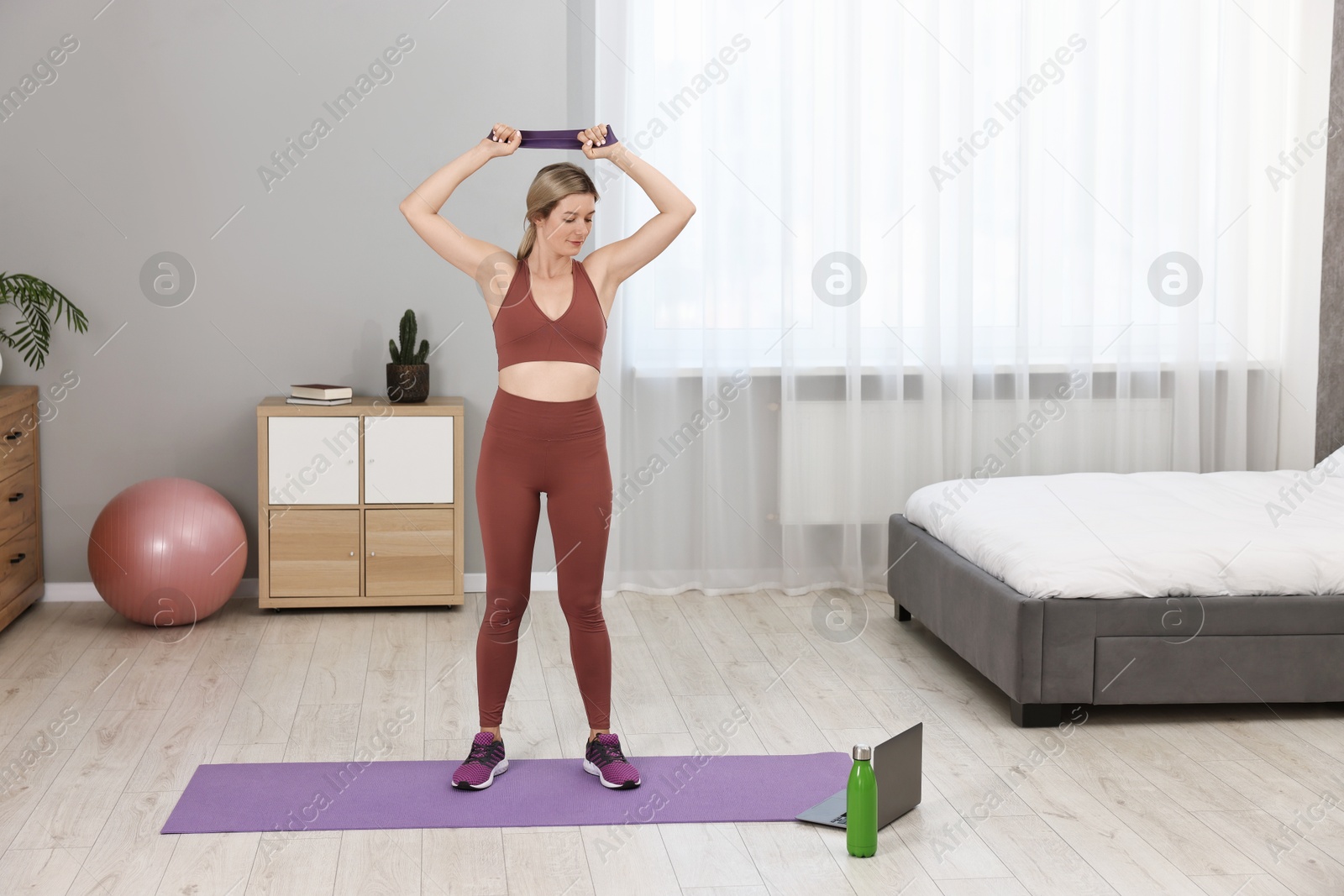 Photo of Online fitness trainer. Woman doing exercise with elastic band near laptop at home