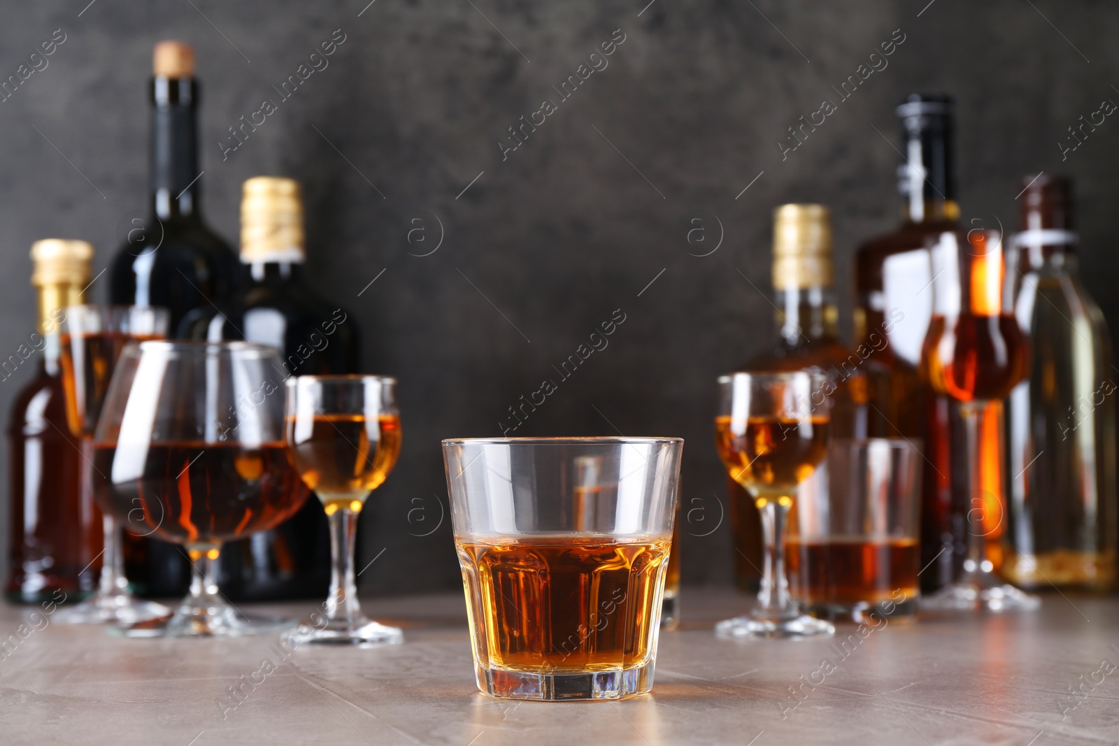 Photo of One liqueur in glass in front of others on grey textured table, closeup