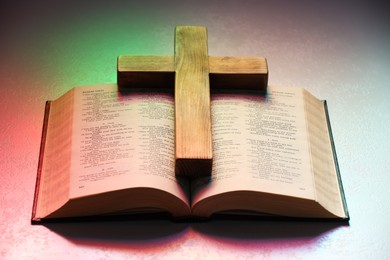 Photo of Wooden cross and Bible on textured table in color lights, closeup. Religion of Christianity