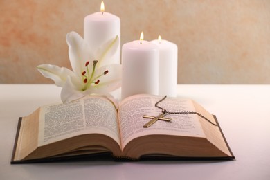 Cross with chain, burning candles, flower and Bible on white table, closeup. Religion of Christianity