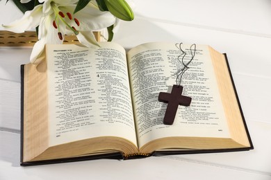 Photo of Cross, Bible and beautiful lily flowers on white wooden table, closeup. Religion of Christianity