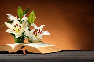 Photo of Bible, cross and lilies on gray table against brown background, space for text. Religion of Christianity