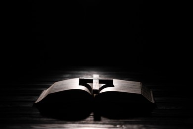 Photo of Bible and cross on table in darkness, space for text. Religion of Christianity