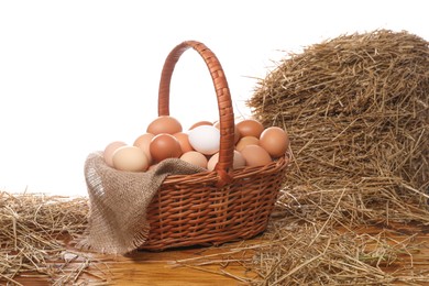 Wicker basket full of chicken eggs and dried straw on wooden table against white background
