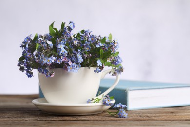 Photo of Beautiful forget-me-not flowers in cup, saucer and book on wooden table against light background, closeup