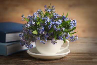 Beautiful forget-me-not flowers in cup, saucer and books on wooden table, closeup