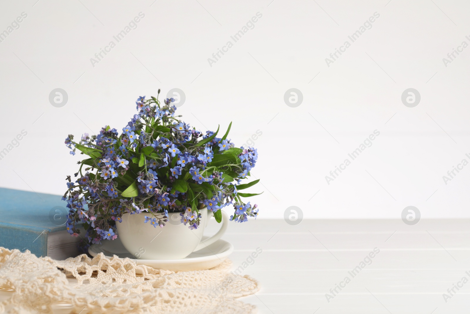 Photo of Beautiful forget-me-not flowers in cup, saucer, crochet tablecloth and book on table against white background, closeup. Space for text