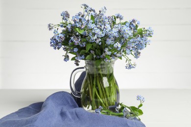 Photo of Bouquet of beautiful forget-me-not flowers in glass jug and blue cloth on white table