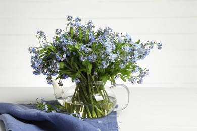 Bouquet of beautiful forget-me-not flowers in glass teapot and blue cloth on white table