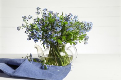 Bouquet of beautiful forget-me-not flowers in glass teapot and blue cloth on white table