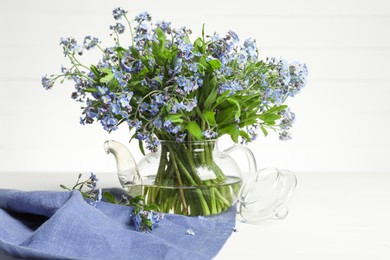 Bouquet of beautiful forget-me-not flowers in glass teapot and blue cloth on white table