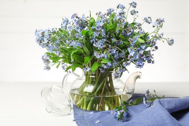 Photo of Bouquet of beautiful forget-me-not flowers in glass teapot and blue cloth on white table
