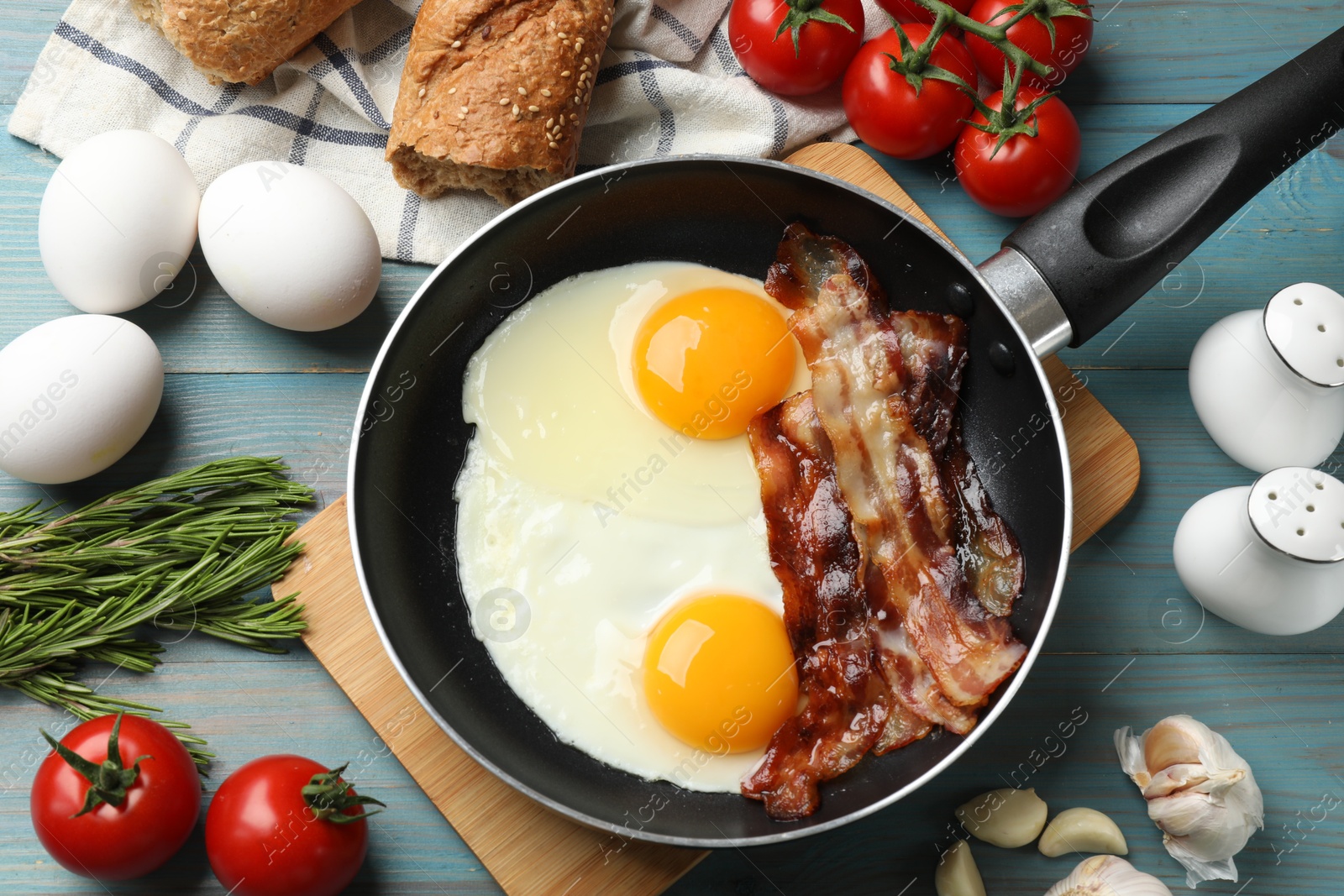 Photo of Tasty bacon and eggs in frying pan among products on light blue table, flat lay