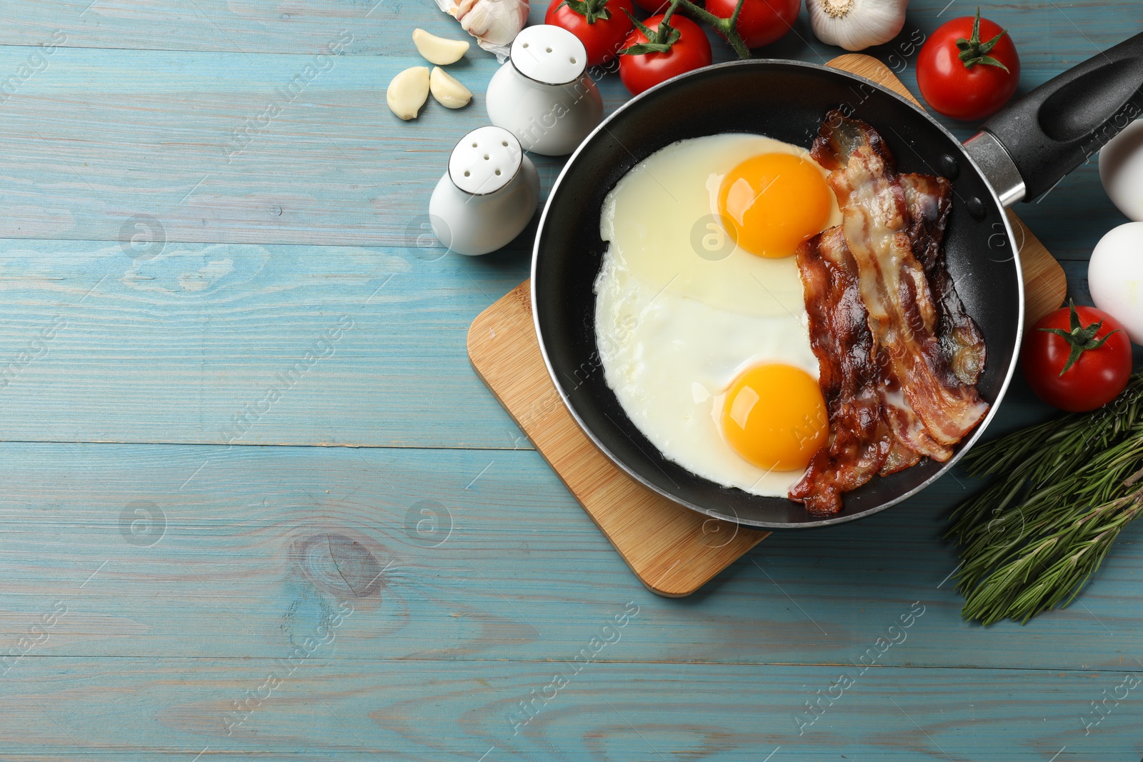 Photo of Tasty bacon and eggs in frying pan among products on light blue table, flat lay. Space for text