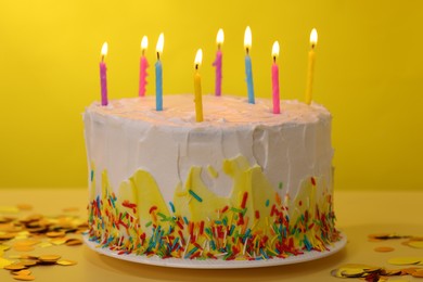 Photo of Tasty Birthday cake with burning candles and confetti on yellow table, closeup