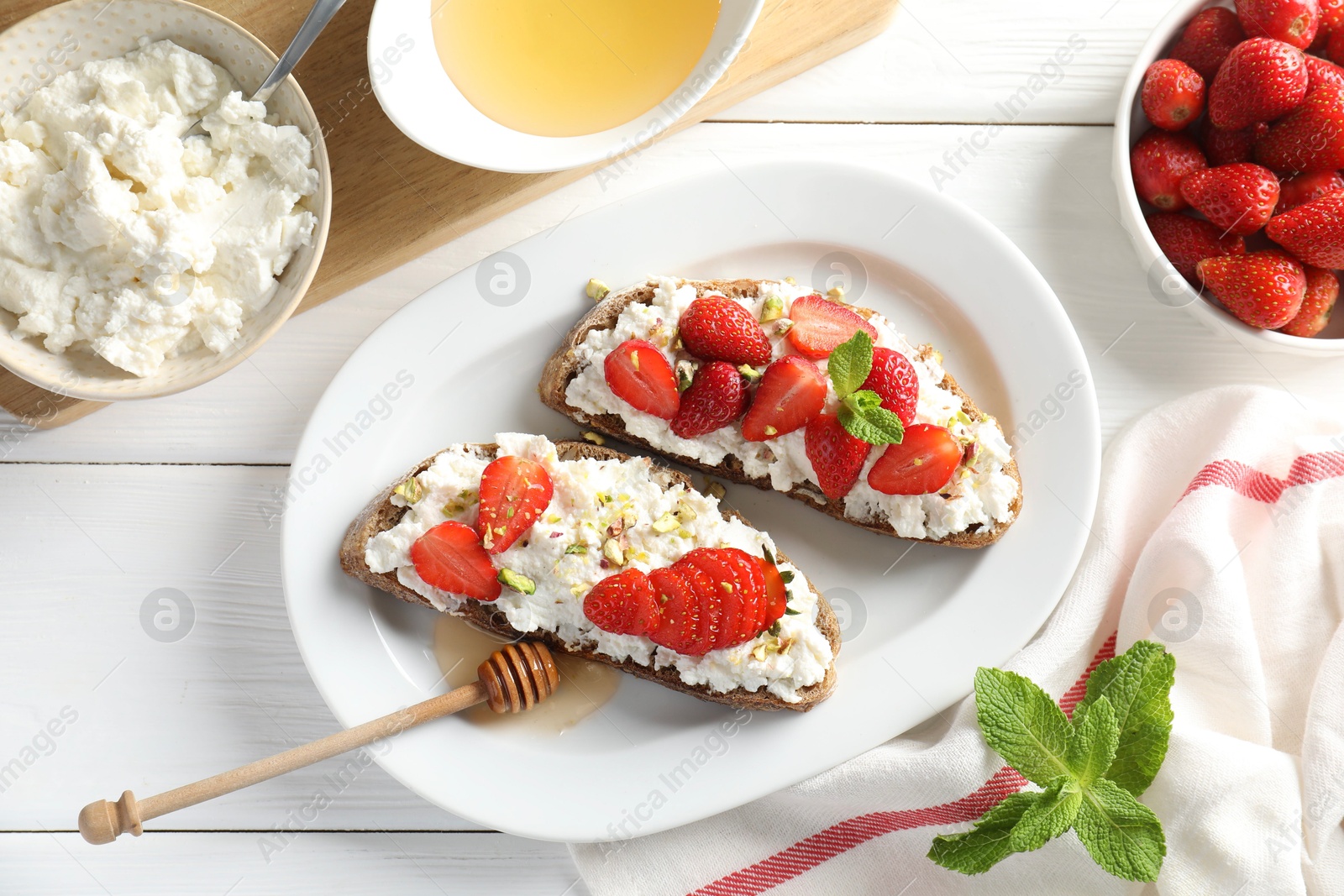 Photo of Delicious ricotta bruschettas with strawberry, mint and pistachios served with honey on white wooden table, flat lay