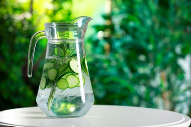 Photo of Refreshing cucumber water with rosemary in jug on white table against blurred green background. Space for text