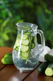 Refreshing cucumber water with rosemary in jug and vegetables on wooden table against blurred green background