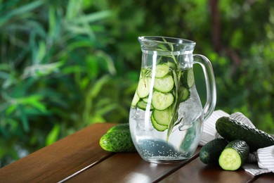 Photo of Refreshing cucumber water with rosemary in jug and vegetables on wooden table against blurred green background. Space for text