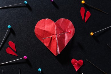 Photo of Halves of torn paper heart connected by sewing pin among other ones on black background, flat lay. Relationship problem concept
