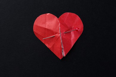 Halves of torn paper heart connected by sewing pin on black background, top view. Relationship problem concept