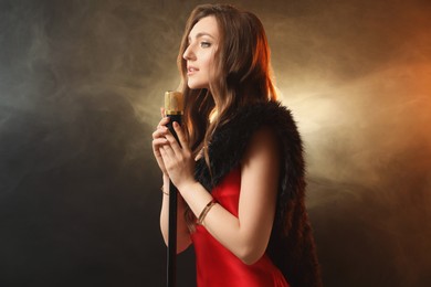 Beautiful young woman with microphone singing on color background with smoke