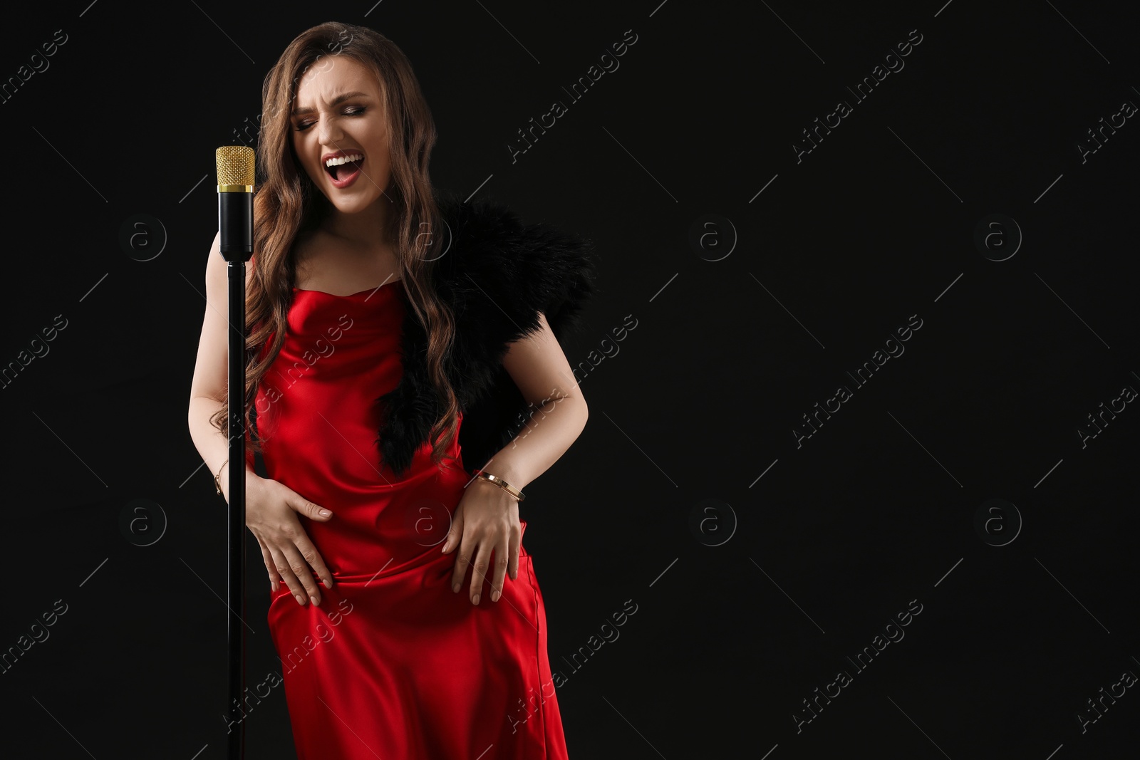 Photo of Beautiful young woman in stylish dress with microphone singing on black background, space for text